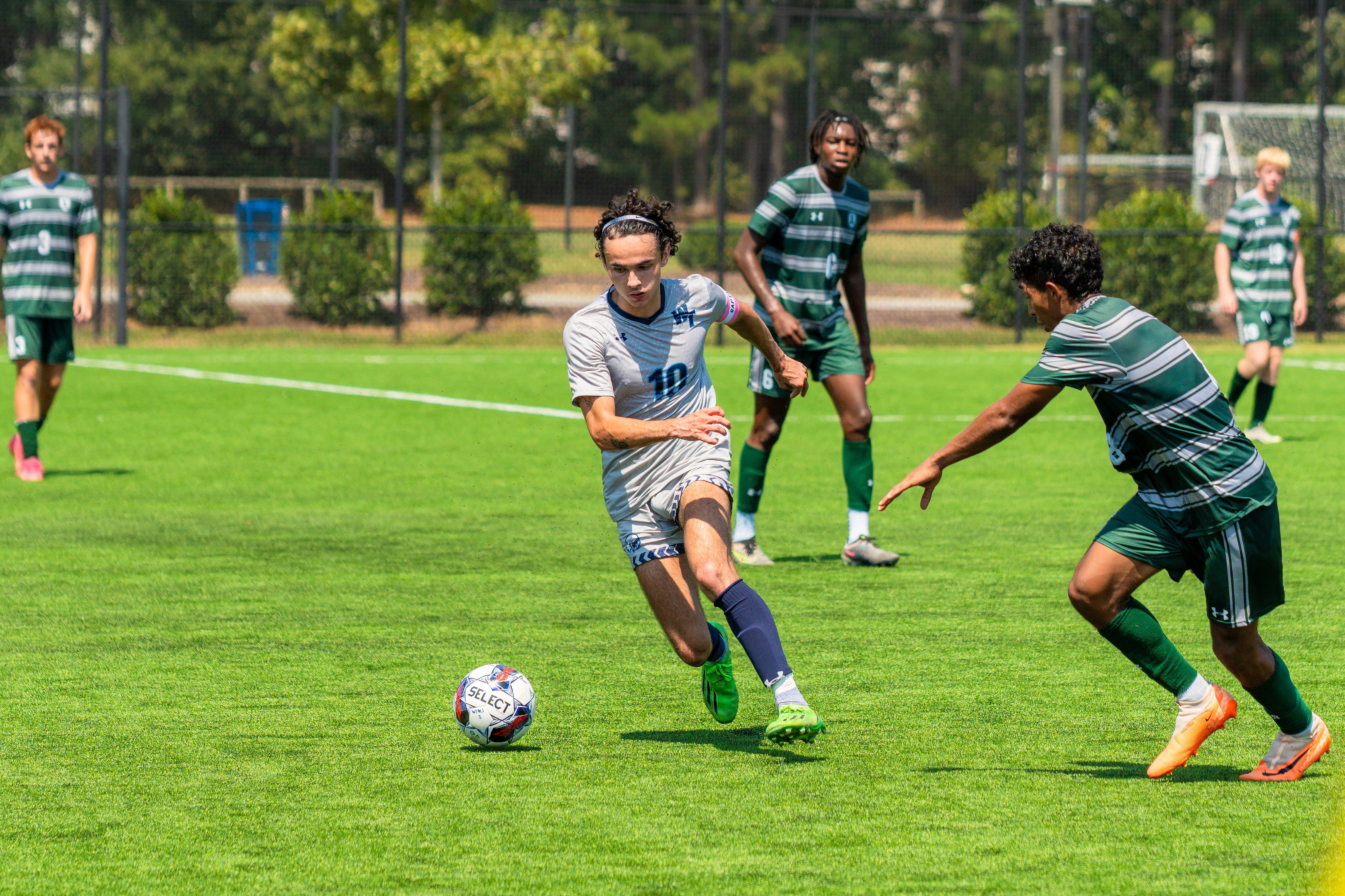 Gruppe junger Männer in Sportkleidung beim Fussballspielen auf einem Rasenfeld mit Bäumen, Maschendrahtzaun und Torpfosten.