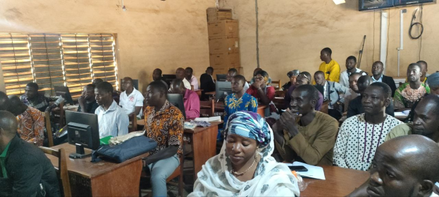 Eine Gruppe von Menschen sitzt an Tischen in einem Klassenzimmer mit Monitoren, Taschen, Büchern, Papieren und Kartons auf den Tischen, einem Fenster auf der linken Seite, einem Fernseher an der Wand im Hintergrund, an einer Techniktrainingssitzung teilnehmend.