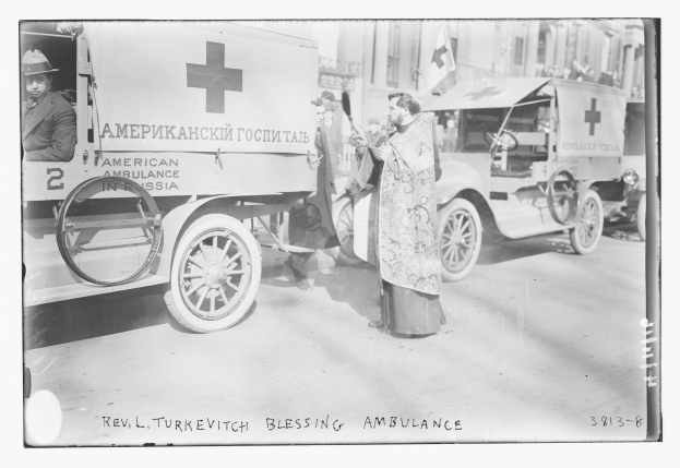 Altes Schwarz-Weiß-Foto eines Mannes neben einem frühen 20. Jahrhundert Krankenwagen auf einer Straße mit anderen Fahrzeugen und Menschen, Gebäuden und einer Flagge im Hintergrund, mit Text unten.