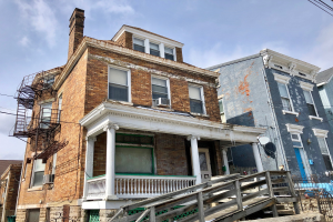 Eine Reihe von baufälligen Häusern an einer Straßenecke, mit zerbrochenen Fenstern, bröckelnden Wänden, Geländern, Stufen, einem Zaun, einer Satellitenschüssel, Kabeln und einem bewölkten Himmel.