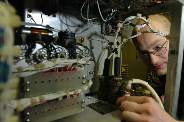 Ein Techniker mit Brille arbeitet an einer Maschine in einem Raum voller elektronischer Geräte und Kabel.