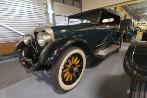 Ein klassisch gestaltetes Oldtimer-Auto mit einer klassischen Design ist in einem Museum ausgestellt, umgeben von anderen Autos, mit einer Wand mit Fensterläden auf der linken Seite und Lampen an der Decke.