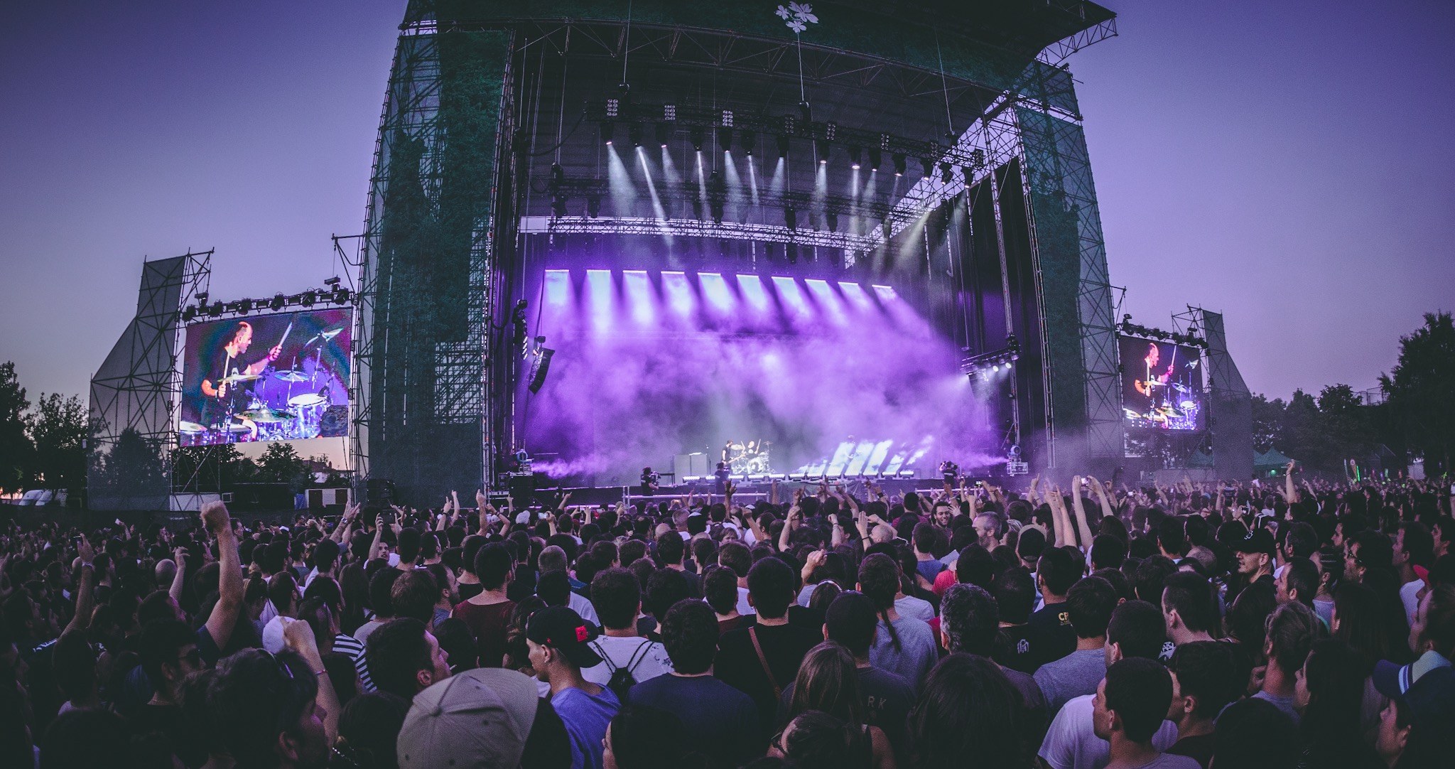 Eine große Menschenmenge bei einem Musikfestival schaut auf der Bühne Performern zu, die von Instrumenten und Bildschirmen umgeben sind, vor einem Gebäude, Bäumen und einem klaren blauen Himmel.