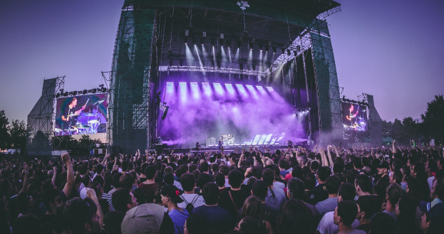 Eine große Menschenmenge bei einem Musikfestival schaut auf der Bühne Performern zu, die von Instrumenten und Bildschirmen umgeben sind, vor einem Gebäude, Bäumen und einem klaren blauen Himmel.