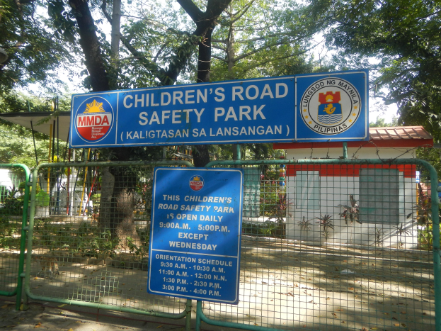 Ein von einem Metallzaun umgebener Kindersicherheitswegpark mit zwei blauen Informationsschildern, einem kleinen Haus mit Dach, Pflanzen, Bäumen und einem bewölkten Himmel.