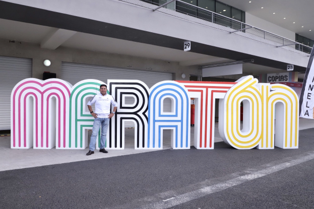 Ein Mann steht vor einem großen "Marathon"-Schild mit einem Banner rechts daneben, mit einem Gebäude mit Lichtern, Geländern und Fensterläden im Hintergrund.