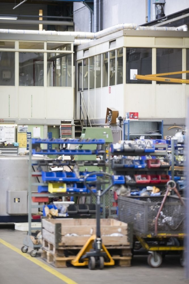 Großer Lagerraum mit verschiedenen Geräten, darunter fahrbare Wagen, Schränke und eine Kiste, mit einer Glaswand und freiliegenden Rohren im Hintergrund.