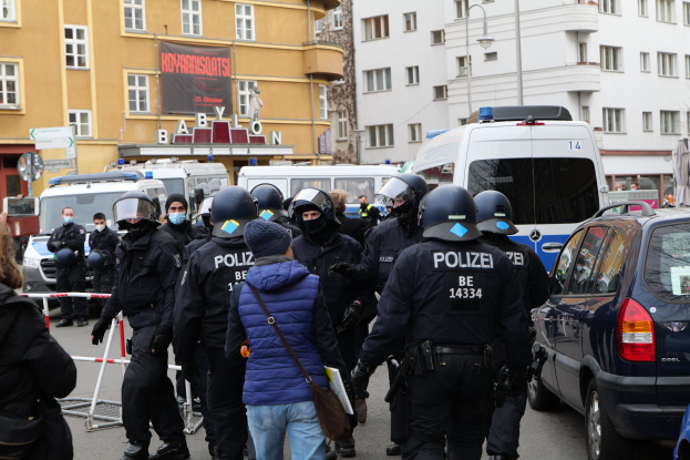 Polizeibeamte in Uniform stehen vor einer Menge mit Helmen und Jacken, mit Fahrzeugen, einer Barriere, Gebäuden, Laternen, Texttafeln, einem Banner und einer Statue im Hintergrund.