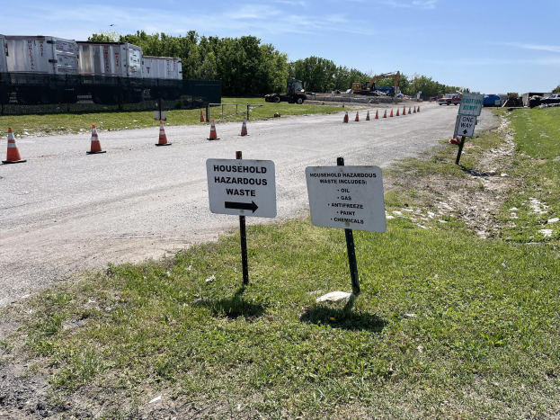 Straßensammelstelle für "Haushaltsgefährliche Abfälle" markiert durch Verkehrskegel, mit Fahrzeugen, Bäumen und einem bewölkten Himmel im Hintergrund.