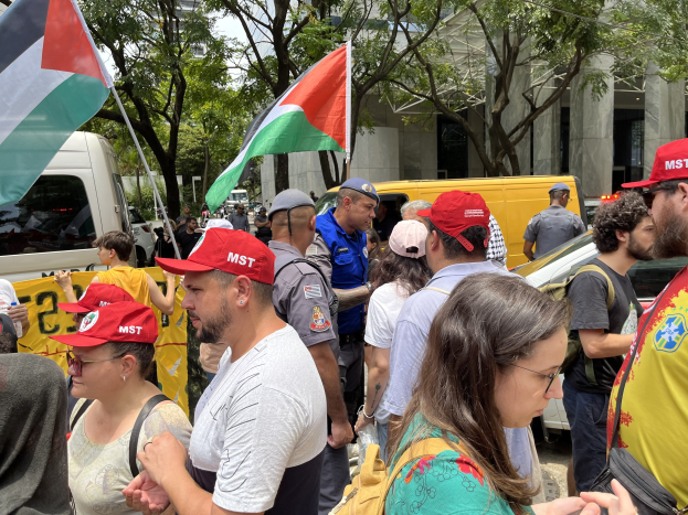 Gruppe von Menschen vor einem Gebäude mit Mützen tragend und Taschen tragend, die palästinensische Flaggen während einer Protestaktion halten, mit Fahrzeugen auf der Straße, Bäumen und einem klaren blauen Himmel im Hintergrund.