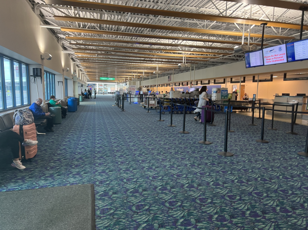 Eine lange Schlange von Menschen, die in einem Flughafen-Terminal warten, mit einigen auf Stühlen sitzend und anderen stehend auf dem Boden, unter Deckenlampen und in der Nähe von Glasfenstern, mit Pfählen und Bildschirmen sichtbar.