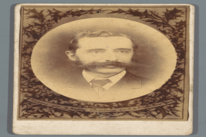 Ein altes Foto eines Mannes mit Schnurrbart und Bart, der direkt in die Kamera schaut und einen ernsten Gesichtsausdruck hat.