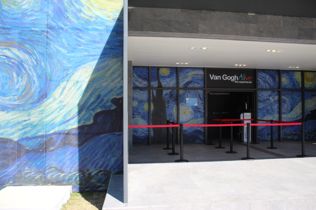 Der Eingang eines Gebäudes zeigt ein großes Wandbild, ein Schild mit der Aufschrift "Van Gogh Alive", Absperrpfosten mit Bändern, Gras auf dem Boden und Lampen an der Decke.
