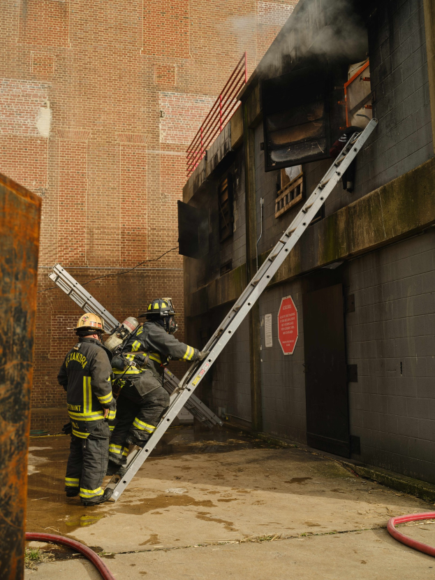 Feuerwehrleute mit Helmen klettern an einer Leiter zu einem raucherfüllten Gebäude hoch, während in der Nähe ein metallisches Objekt und Bodenschläuche zu sehen sind.