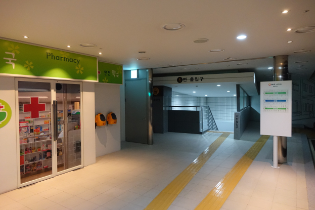 Apothekeneingang mit einem "Apotheke"-Schild auf der linken Seite, einer Tafel mit Text auf der rechten Seite, beleuchteten Lichtern oben und Hintergrundobjekten.