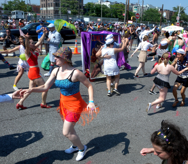 Eine Gruppe von Menschen, die eine Stra√e entlangmarschieren, einige mit Mötzen und Schirmen, andere tanzend, mit einer Fahne, Verkehrskegeln, Fahrzeugen, Lichtmästen, Schildern, Bäumen, Gebäuden und einem klaren blauen Himmel im Hintergrund.
