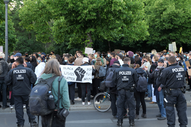 Eine große Gruppe von Menschen mit Schildern und Mützen und Taschen steht an einer Straße, mit einem Fahrrad im Vordergrund und Bäumen und einem Pfahl im Hintergrund, was auf eine Black Lives Matter Demonstration in Berlin hinweist.