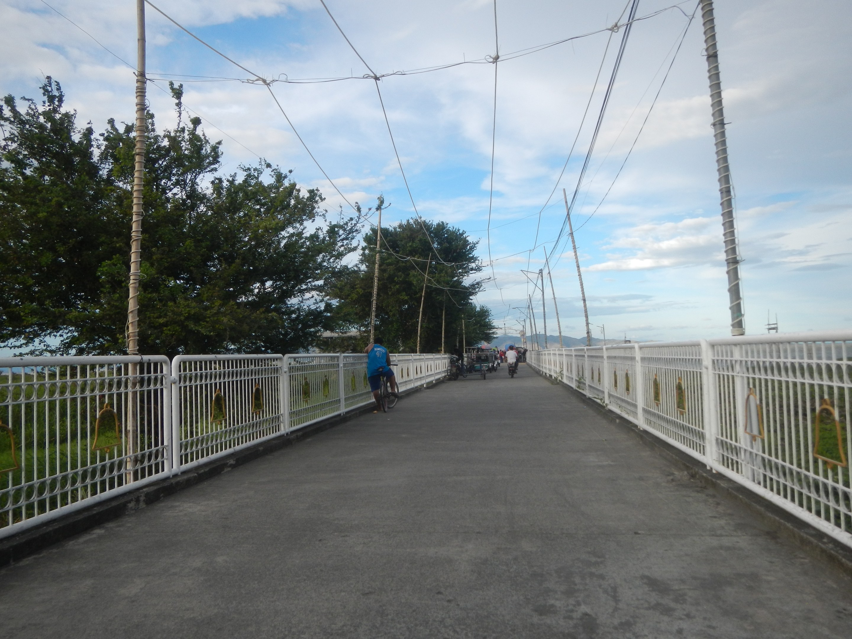 Eine Fußgängerbrücke mit Radfahrern, Geländern, Strommasten mit Kabeln, Bäumen und einem bewölkten Himmel im Hintergrund.