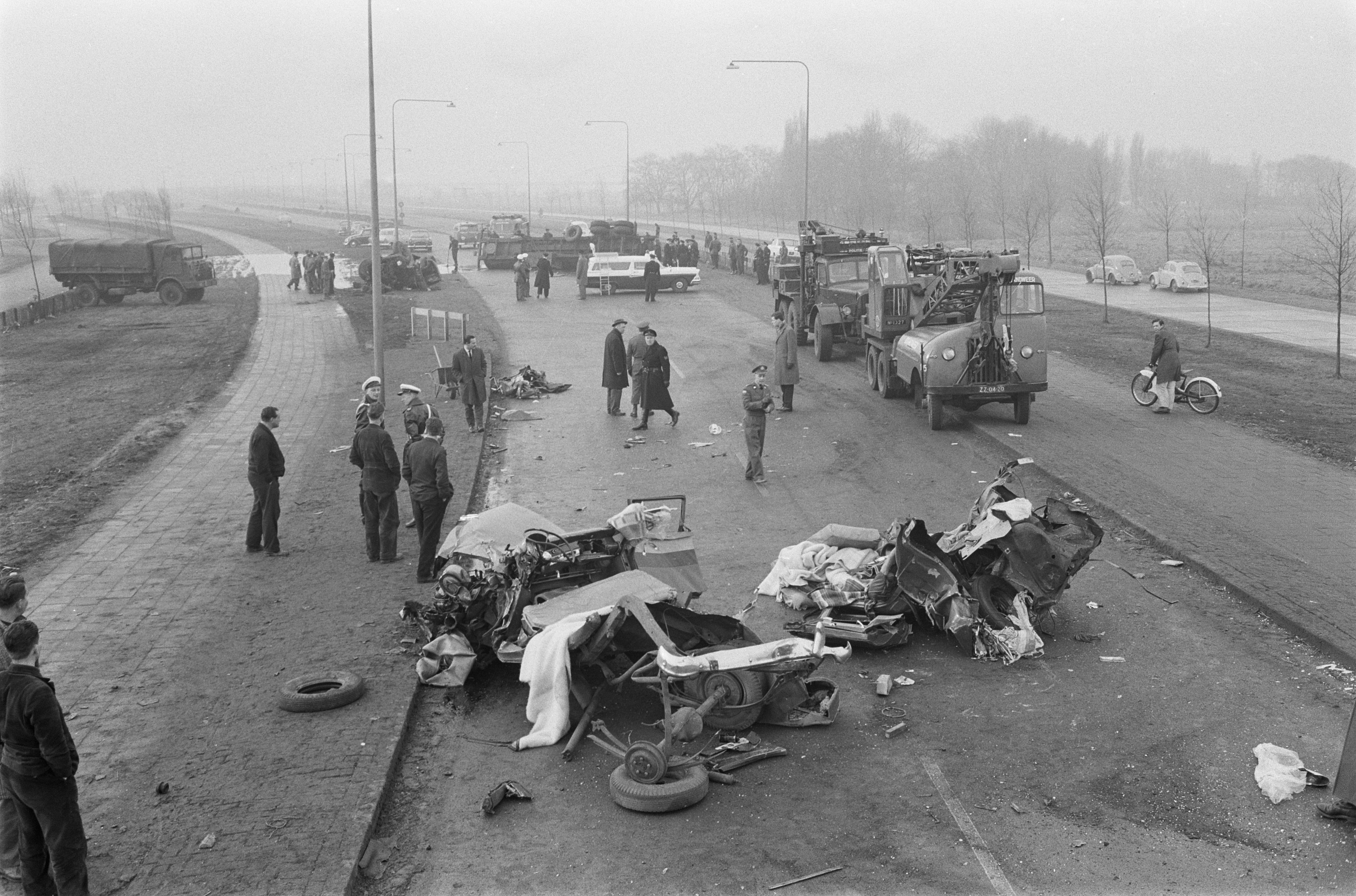 Schwarze und Weiße Szenerie eines Autounfalls am Straßenrand mit mehreren Fahrzeugen, einer Gruppe von Menschen in der Nähe, Laternen, Bäumen und Himmel im Hintergrund.