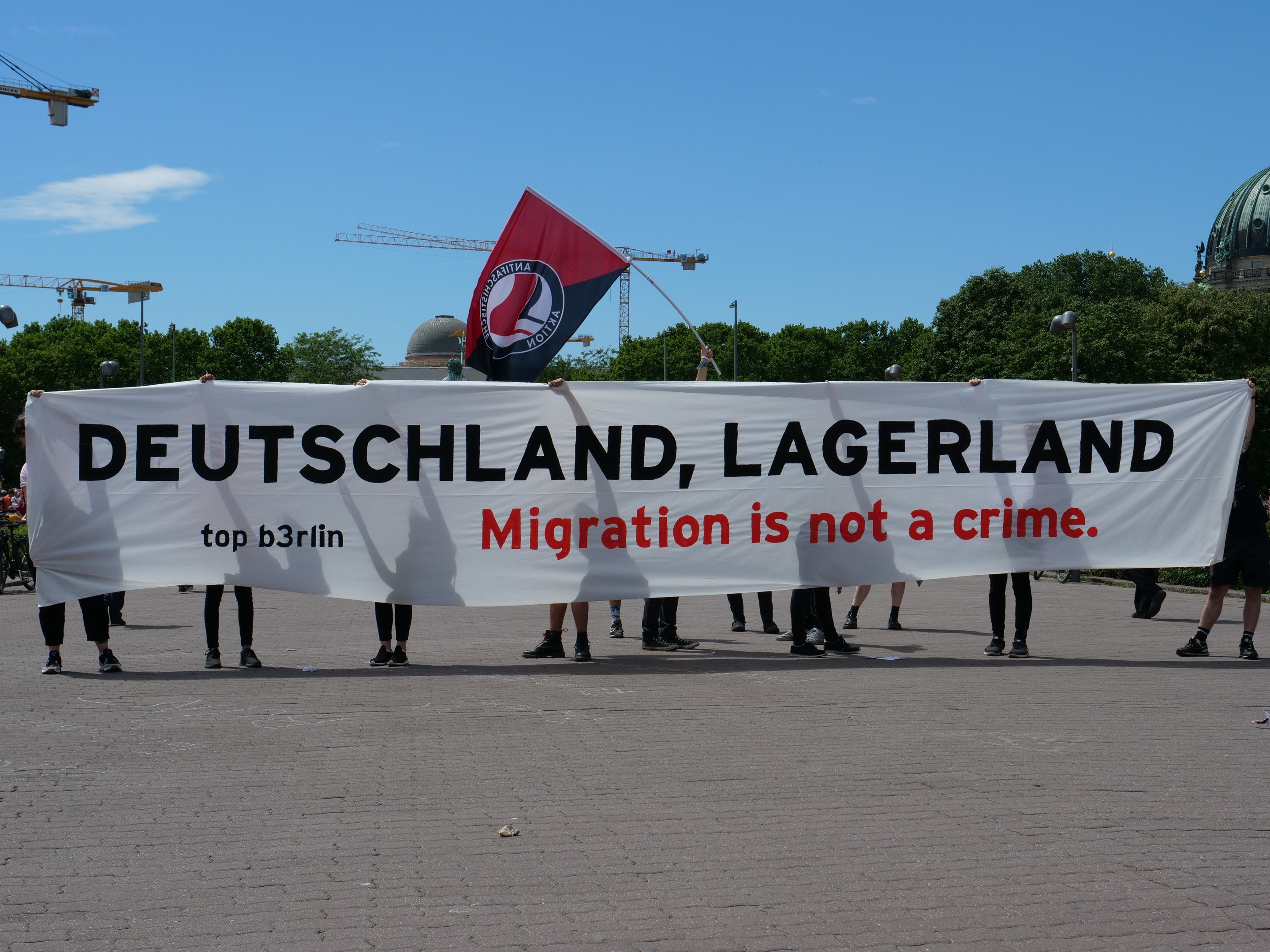 Menschen mit einem Banner, auf dem "Deutschland, Lagerland Migration ist kein Verbrechen" und einer Flagge, mit Straßenlaternen, Bäumen, Kränen, einem Gebäude und einem bewölkten Himmel im Hintergrund.