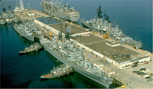 Luftaufnahme eines großen Kriegsschiffs, das in einem Hafen liegt und von anderen Schiffen umgeben ist, mit Fahrzeugen auf der rechten Seite und Text unten.