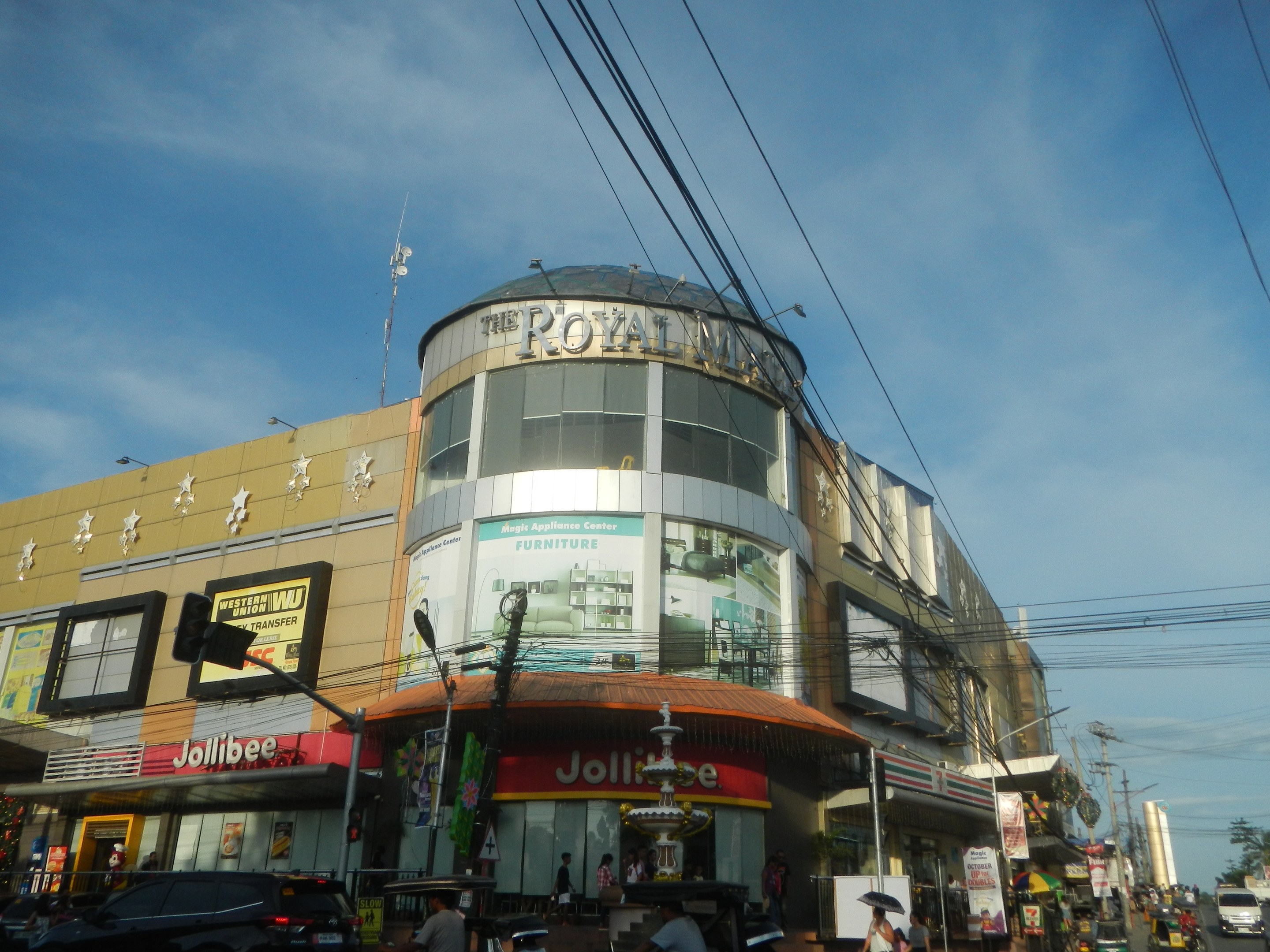 Eine belebte Stadtstraße mit fahrenden Fahrzeugen, Fußgängern auf dem Gehweg und einem großen Gebäude mit der Aufschrift "Royal Mall" im Hintergrund, umgeben von parkenden Autos, Bäumen, Strommasten und Drähten unter dem Himmel.