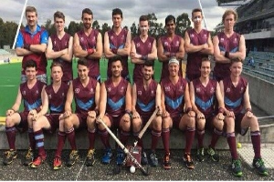 Eine Gruppe von Männern, die auf einem Feld mit Hockeystöcken feiern, mit einem Stadion und Zuschauern im Hintergrund unter einem klaren blauen Himmel.
