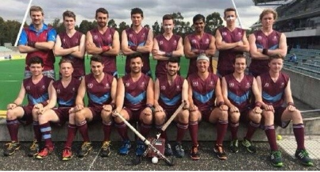 Eine Gruppe von Männern, die auf einem Feld mit Hockeystöcken feiern, mit einem Stadion und Zuschauern im Hintergrund unter einem klaren blauen Himmel.