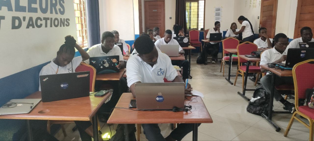 Eine Gruppe von Schülern in einem Klassenzimmer mit Laptops, Taschen auf dem Boden und einer Wand mit Text, die auf einen Online-Kurs in Ghana hinweist, mit Fenstern und Türen im Hintergrund.