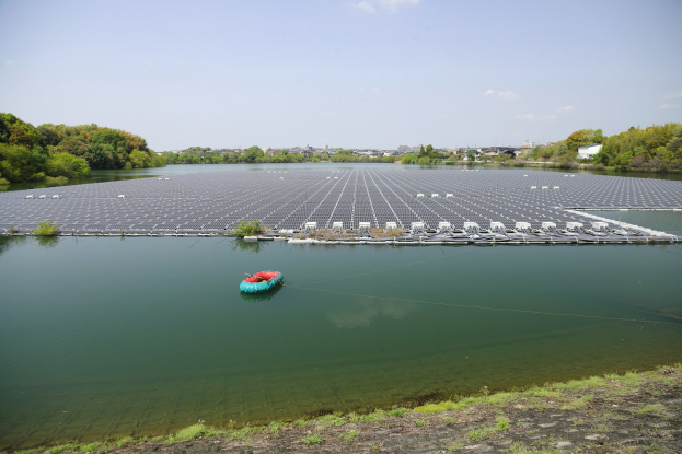 Ein kleines Boot schwimmt auf einem Gewässer, umgeben von grünem Gras und Bäumen, mit Gebäuden und einem klaren blauen Himmel im Hintergrund und Solarmodulen auf der Wasseroberfläche.