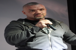 Ein Mann in einer schwarzen Kapuzenjacke und einem schwarzen Anzug mit einem weißen Hemd hält ein Mikrofon und steht selbstbewusst vor einer Wand, wobei er wie ein Komiker mit zurückgekämmtem Haar wirkt.