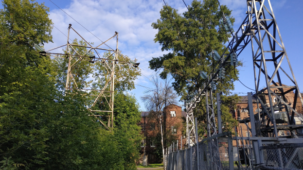 Gro├če Stromleitung durch einen Wald mit umliegenden B├Ąumen, Masten, Dr├Ąhten und einem Zaun, mit im Hintergrund sichtbaren Geb├Ąuden unter einem bew├Âlktem Himmel.