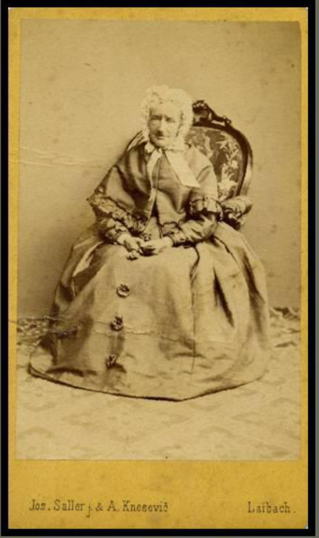 Ein altes Foto einer Frau mit sanftem Gesichtsausdruck, die ein helles Kleid und einen gemusterten Schal trägt und in einem Stuhl sitzt, mit ihren Händen auf dem Schoß ruhend, ihr Haar ordentlich zurückgekämmt, in einem Foto Rahmen ausgestellt.