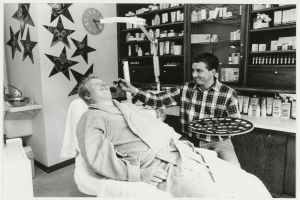 Ein Mann lächelt in einem Zahnarztstuhl, während ein anderer Mann neben ihm steht und ein Tablett hält, mit einem Waschbecken, Uhr und Regalen mit Flaschen im Hintergrund.