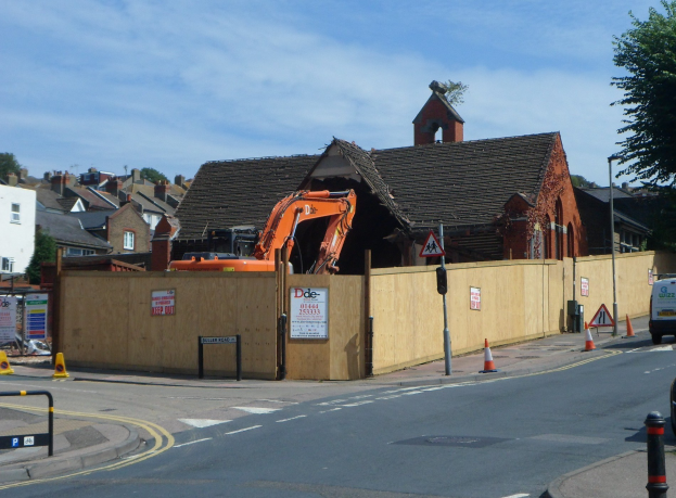 Straße mit Fahrzeugen, Verkehrskegeln, Pfählen, Schildern und einem Zaun; Bagger und teilweise abgerissene Gebäude am Straßenrand; Gebäude, Bäume, Wolken und Himmel im Hintergrund.