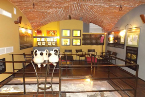 Ein Museumsraum voller Tische, Stühle, eines Geländers, Vasen, Texttafeln, Wandrahmen, Lampen und anderer Ausstellungsstücke.