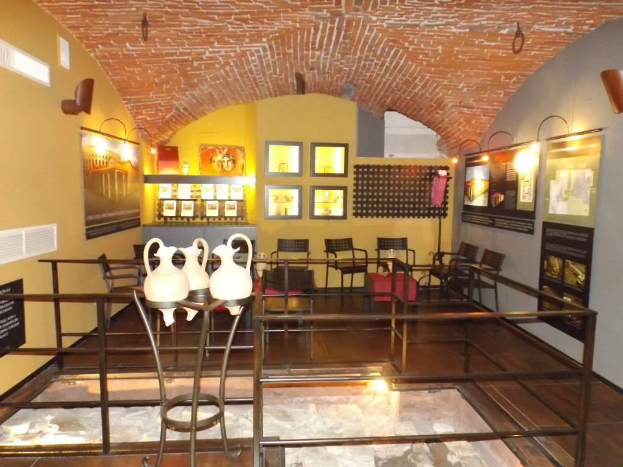 Ein Museumsraum voller Tische, Stühle, eines Geländers, Vasen, Texttafeln, Wandrahmen, Lampen und anderer Ausstellungsstücke.