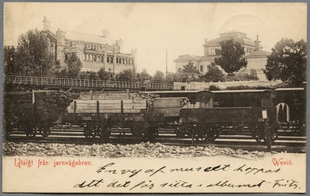 Altes Schwarz-Wei├č-Foto einer Dampflokomotive auf Eisenbahnschienen mit einer ├Überquerung und Geb├Ąuden im Hintergrund, umgeben von B├Ąumen und Text am unteren Rand.