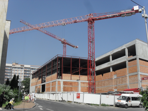 Eine Baustelle mit einem großen Gebäude im Bau, umgeben von Kränen, Fahrzeugen, Menschen, die gehen, Bäumen, Pflanzen, Gras, Schildern, Straßenlaternen und einem klaren blauen Himmel.
