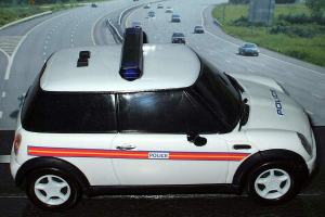 Ein weißer Polizeiwagen steht im Vordergrund mit mehreren Fahrzeugen, die dahinter auf der Straße fahren, umgeben von Gras und Bäumen auf beiden Seiten.