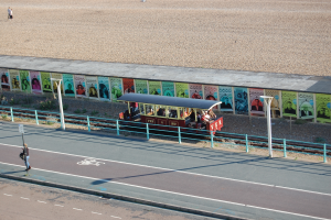 Ein kleiner Zug mit Passagieren fährt auf Schienen neben einem Strand entlang, mit einer Person zu Fuß auf einem Fußweg links, einer Wand mit Plakaten im Hintergrund und sichtbarem Boden oben im Bild.