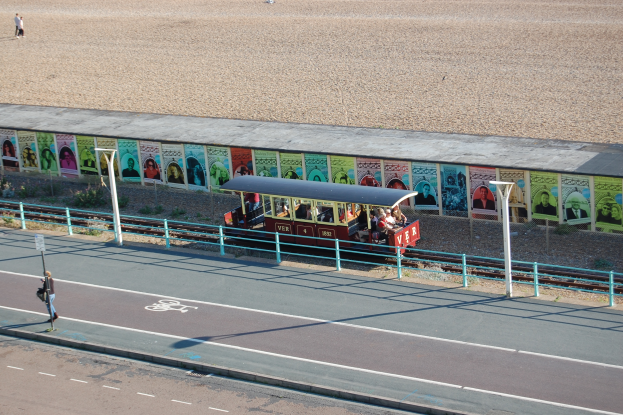 Ein kleiner Zug mit Passagieren fährt auf Schienen neben einem Strand entlang, mit einer Person zu Fuß auf einem Fußweg links, einer Wand mit Plakaten im Hintergrund und sichtbarem Boden oben im Bild.