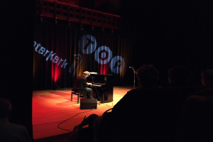 Ein Mann in einer Mütze sitzt an einem Piano auf einer schwach beleuchteten Bühne, mit einem Mikrofonständer rechts daneben und einem Vorhang mit Text im Hintergrund.