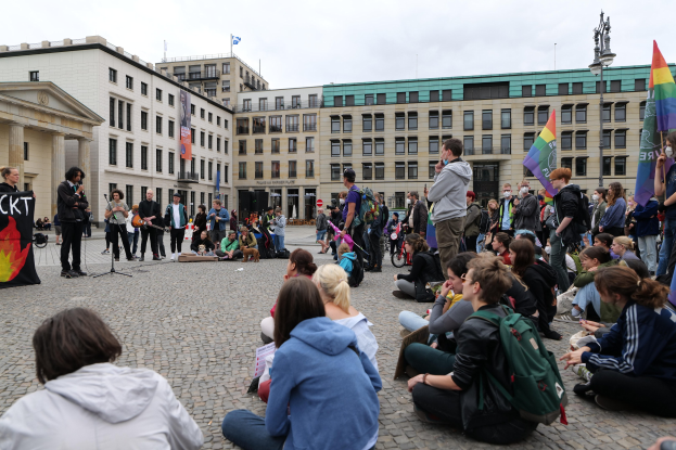 Eine Gruppe von Menschen, die auf dem Boden vor einer Menge sitzen, die Fahnen und Transparente schwenkt, vor einer Statue, Gebäuden und einem bewölktem Himmel bei einer Anti-Schwulen-Demo in Berlin.