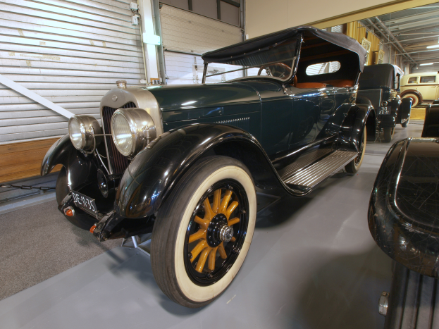 Ein klassisch gestaltetes Oldtimer-Auto mit einer klassischen Design ist in einem Museum ausgestellt, umgeben von anderen Autos, mit einer Wand mit Fensterläden auf der linken Seite und Lampen an der Decke.