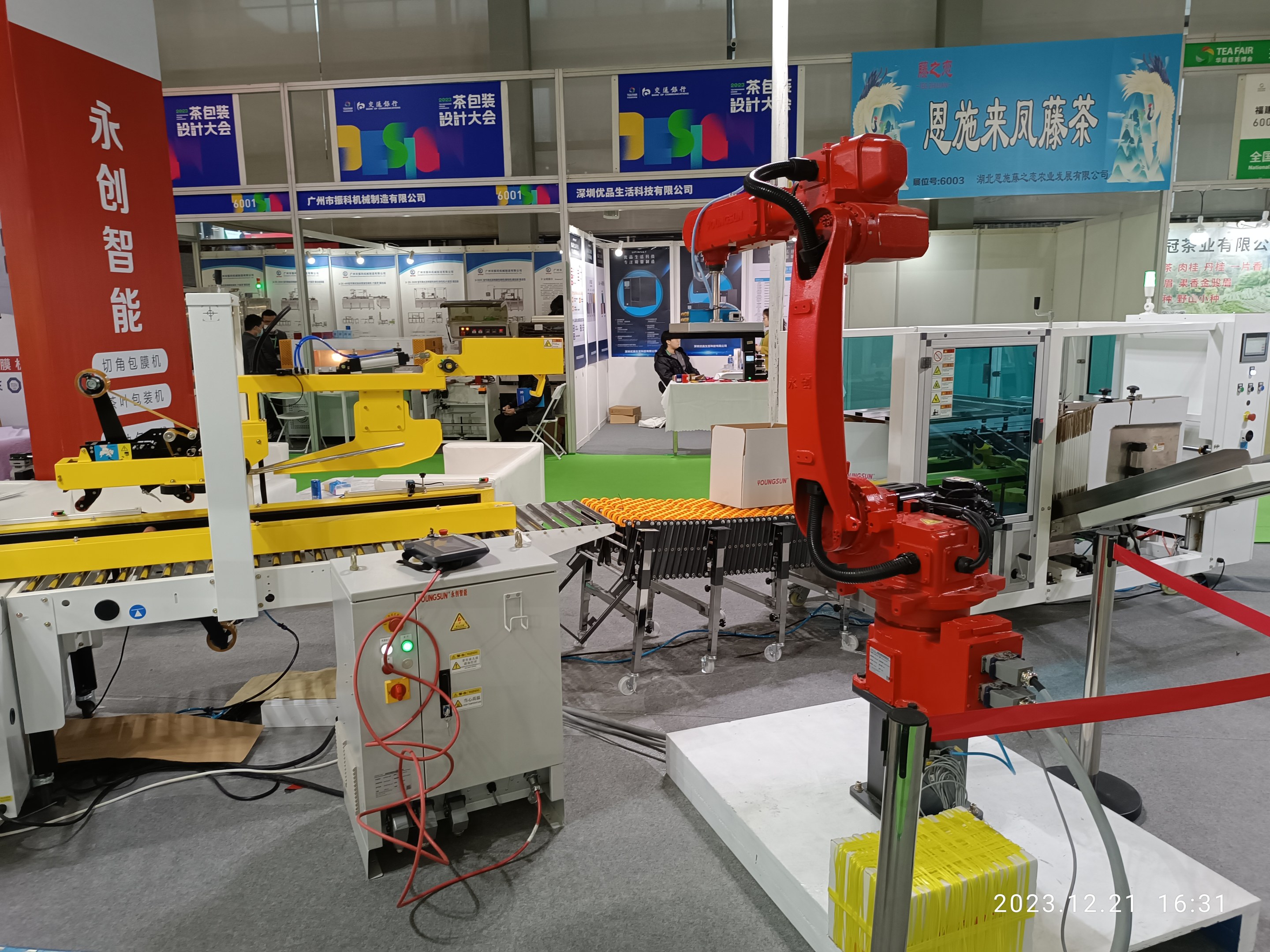 Gruppe von Robotern auf einem Tisch in einer Fabrikumgebung umgeben von Maschinen, Kabeln, textbedruckten Platinen und beleuchteten Komponenten, mit Menschen im Hintergrund.