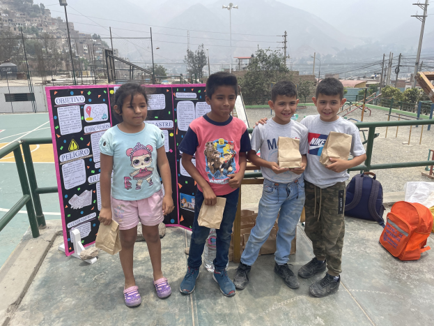 Kinder auf einem Basketballfeld mit Papiertüten stehend, mit Gegenständen auf der rechten Seite und einer Tafel mit Text auf der linken Seite, vor einem Hintergrund aus Zäunen, Pfosten, Straßenlaternen, Bäumen, Gebäuden, Hügeln und dem Himmel.