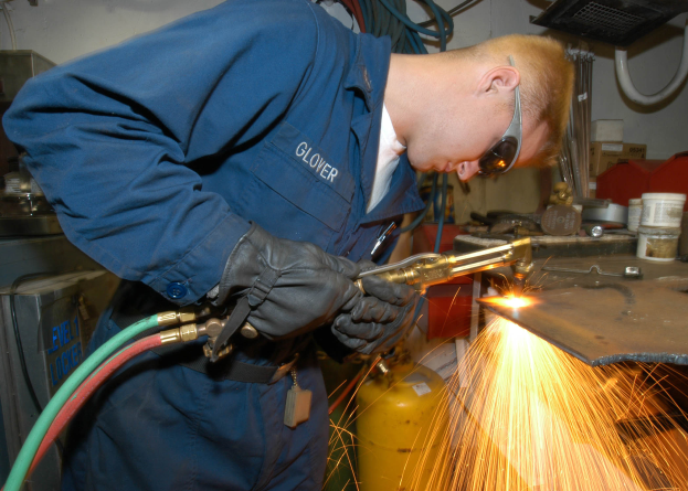 Ein Schweißer in Schutzbrille und Handschuhen arbeitet an einem Metallstück und erzeugt Funken, mit Rohren, Kisten und anderen Gegenständen im Hintergrund.