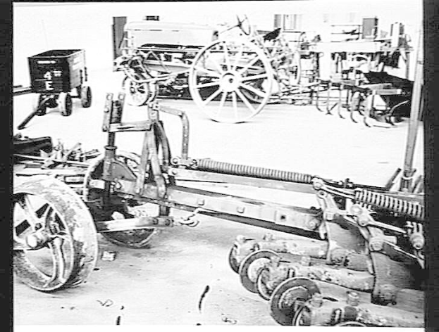 Schwarz-weißes Foto eines Traktors in einer Fabrik, mit einer Egge im Vordergrund und einer Wand im Hintergrund, eingerahmt von einem Rahmen.