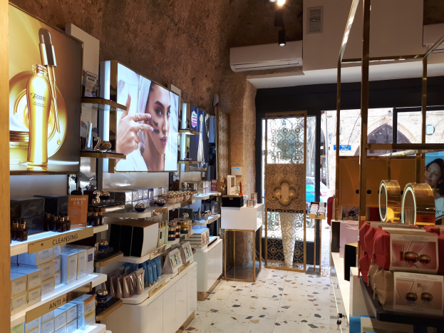 Innenseite eines Kosmetikladens mit verschiedenen Produkten auf Gestellen, Bildschirmen an den Wänden, Deckenleuchten, einer Klimaanlage und Glastüren, die eine Person, ein Auto und ein Gebäude zeigen.